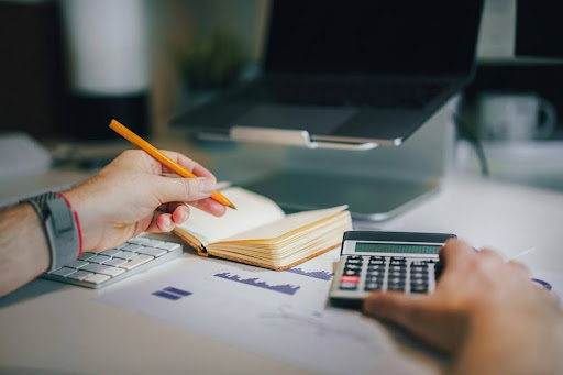 A person making notes in a small notebook beside a calculator, charts and a laptop