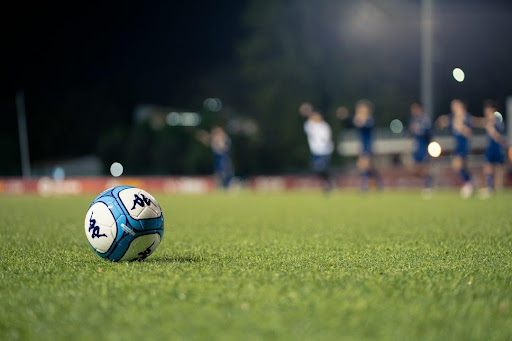 A soccer ball on a grass pitch at night, with players and floodlights blurred in the background